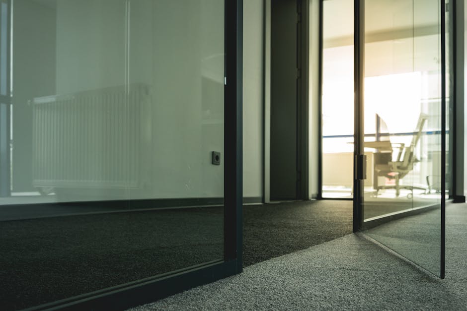 Interior view of a modern office space featuring glass partitions with black metal frames, a textured dark gray carpeted floor, and a softly illuminated area with natural light coming through large windows. A visible office chair and desk can be seen in the background beyond the glass, highlighting the clean and well-maintained environment. Pimlico Carpet Cleaning's surface cleaning and deep cleaning services ensure hygienic, spotless commercial office spaces, as demonstrated in this contemporary setting.