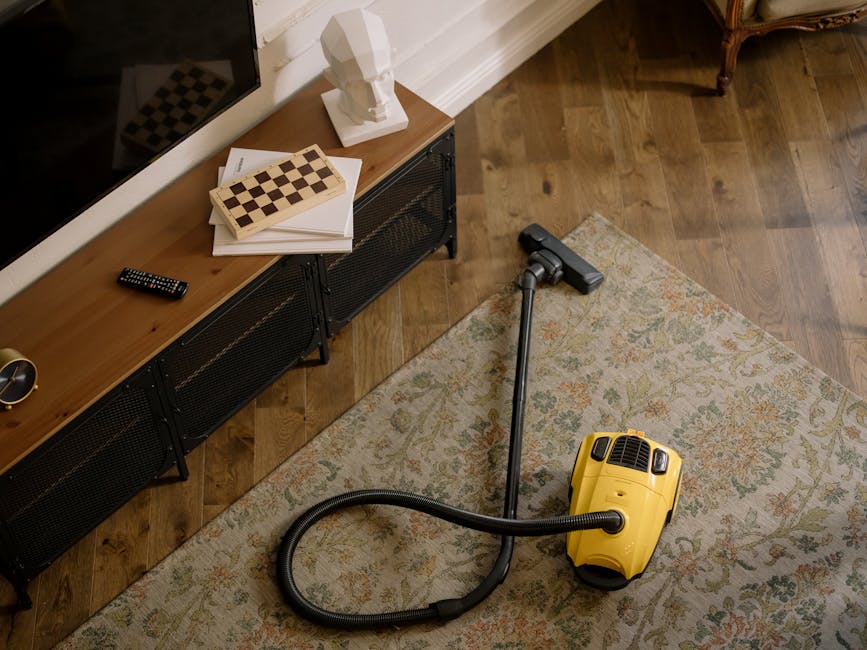 A person dressed in a beige jumpsuit is operating a small, portable vacuum cleaner on a dark carpeted floor in an indoor setting. The background features a black wall with a large white panel or proje