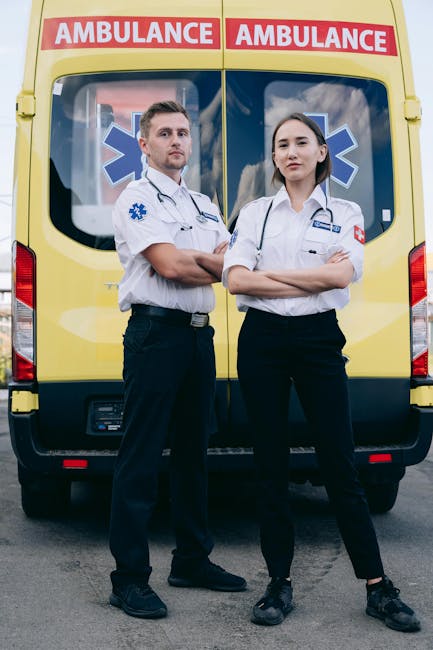 A pair of paramedics, a man and a woman dressed in white medical uniforms with stethoscopes around their necks, standing confidently with crossed arms in front of a yellow ambulance with red and white emergency markings and blue medical symbols on the back windows, outdoors on a paved surface with a clear sky background.