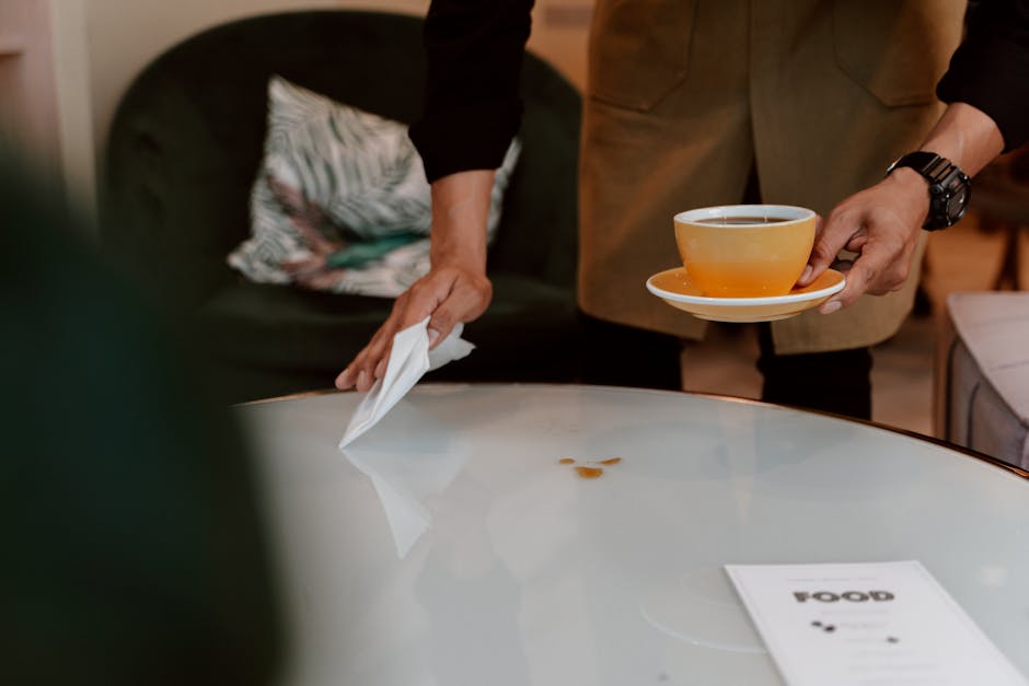 A person wearing a brown blazer is seen cleaning a white, glossy round table in a room with warm lighting, possibly a hotel lobby or office reception area. The individual is holding a white cloth in one hand and a beige cup filled with a hot beverage on a matching saucer in the other. There are small coffee stains on the table surface, which the person appears to be addressing. In the background, a black sofa with a patterned cushion is visible, contributing to a cozy and upscale environment. The scene emphasizes surface cleaning and maintenance to preserve cleanliness and hygiene, aligning with Pimlico Carpet Cleaning's emphasis on professional cleaning services. The setting suggests a focus on deep cleaning or surface sanitisation for public or commercial spaces.