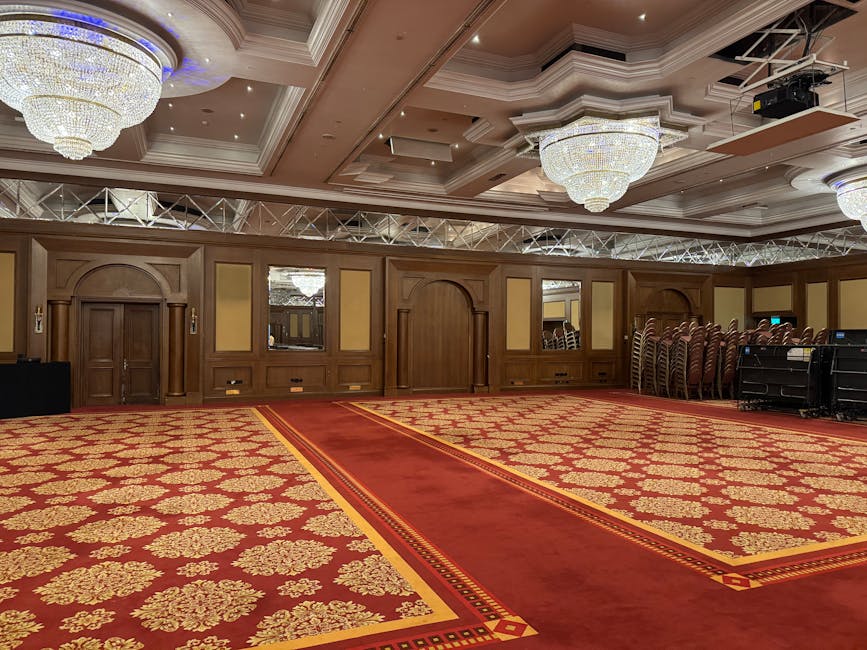 An expansive meeting or event room with a patterned red and gold carpet featuring floral motifs, illuminated by large ornate chandeliers hanging from a detailed white ceiling with recessed lighting. The room has wooden-paneled walls with arched doorways and mirrored panels, and a stack of chairs is neatly arranged along one side, indicating preparation for a gathering. Visible equipment, possibly audio-visual, is situated near the chairs. The space appears clean and well-maintained, suitable for hosting formal events or conferences, with the room managed by Pimlico Carpet Cleaning as part of their surface cleaning and deep cleaning services.