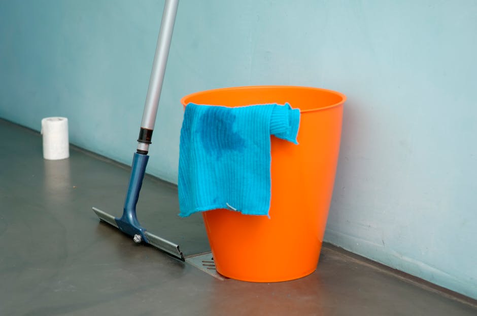 Close-up of an orange plastic bucket placed on a smooth grey floor next to a wall, with a blue cleaning cloth draped over the edge of the bucket, showing some dampness. A mop with a silver handle and dark blue base leans against the bucket, positioned near a floor drain. In the background, a roll of white paper towels is visible against the wall, indicating a cleaning or maintenance setting at the Tate Britain event spill cleanup. The scene depicts surface cleaning tools and damp cloths, emphasizing hygiene and dedicated cleaning processes in a modern, well-lit interior space. Pimlico Carpet Cleaning specializes in deep cleaning and sanitisation, ensuring surfaces are thoroughly maintained.